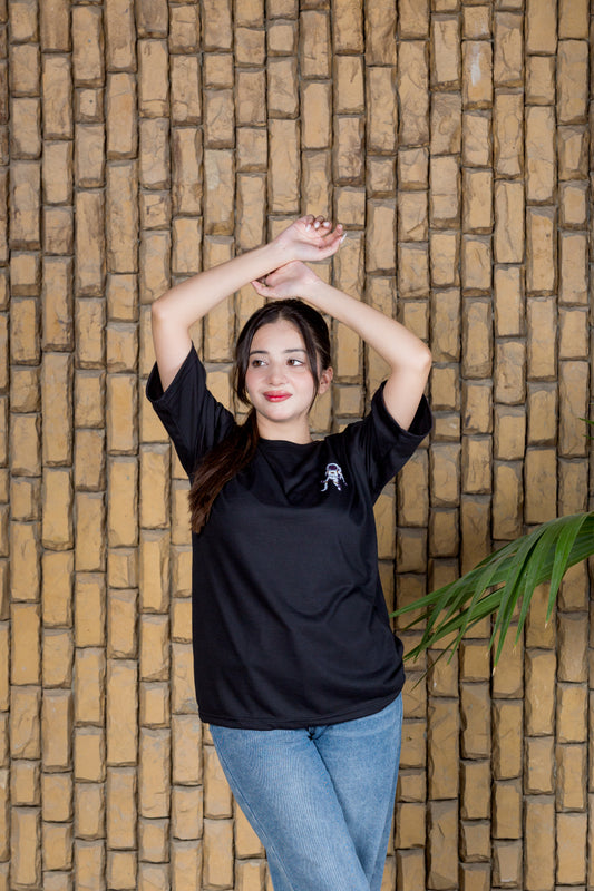 Person wearing a black t-shirt with a logo against a wooden paneled wall.